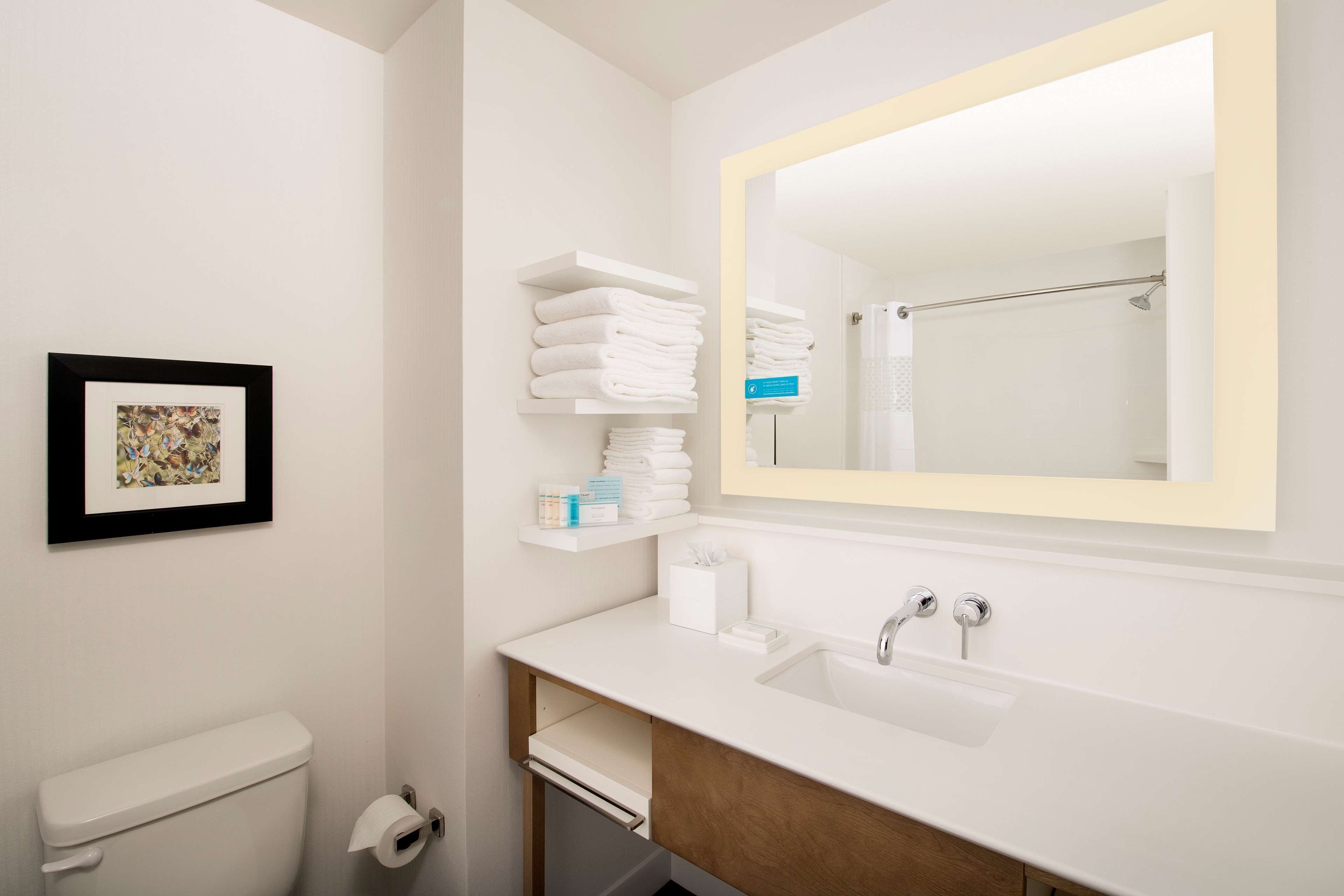 Un baño moderno con inodoro, lavamanos y un espejo grande. Hay repisas con toallas dobladas y artículos de aseo.