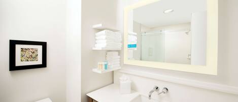 A modern bathroom with a white sink, a large mirror, and a toilet. There are shelves with folded towels and toiletries.