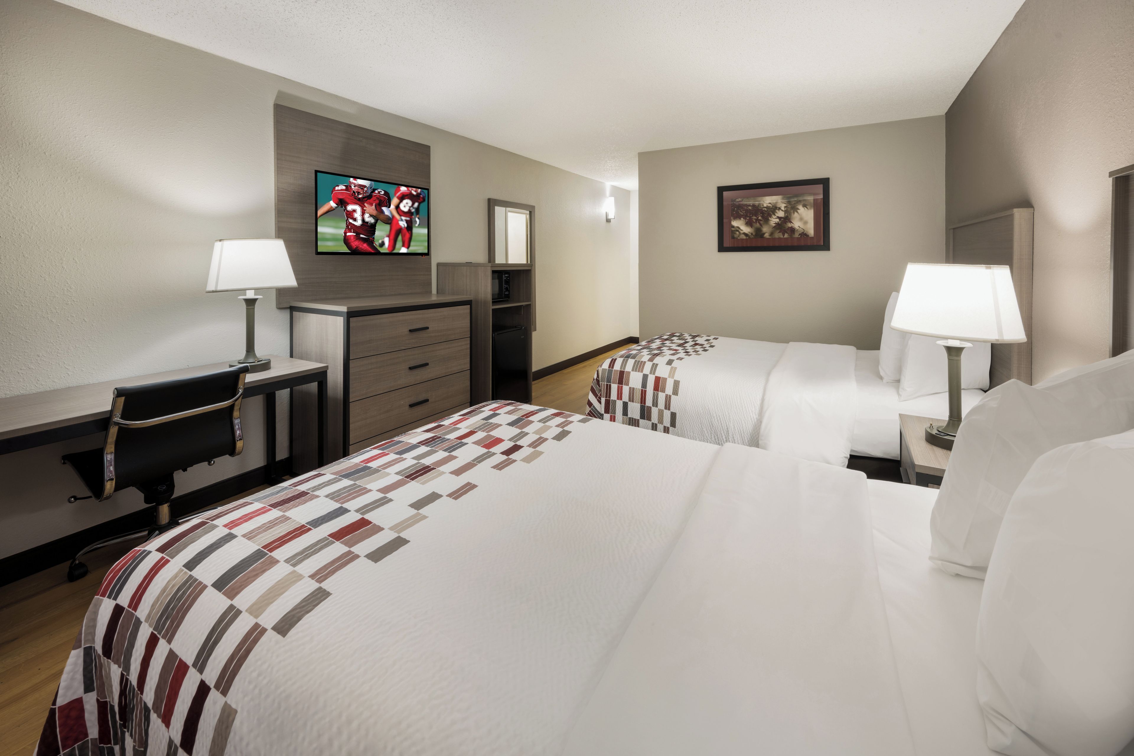 A hotel room with two beds, a desk, a dresser, a TV, and a painting on the wall.