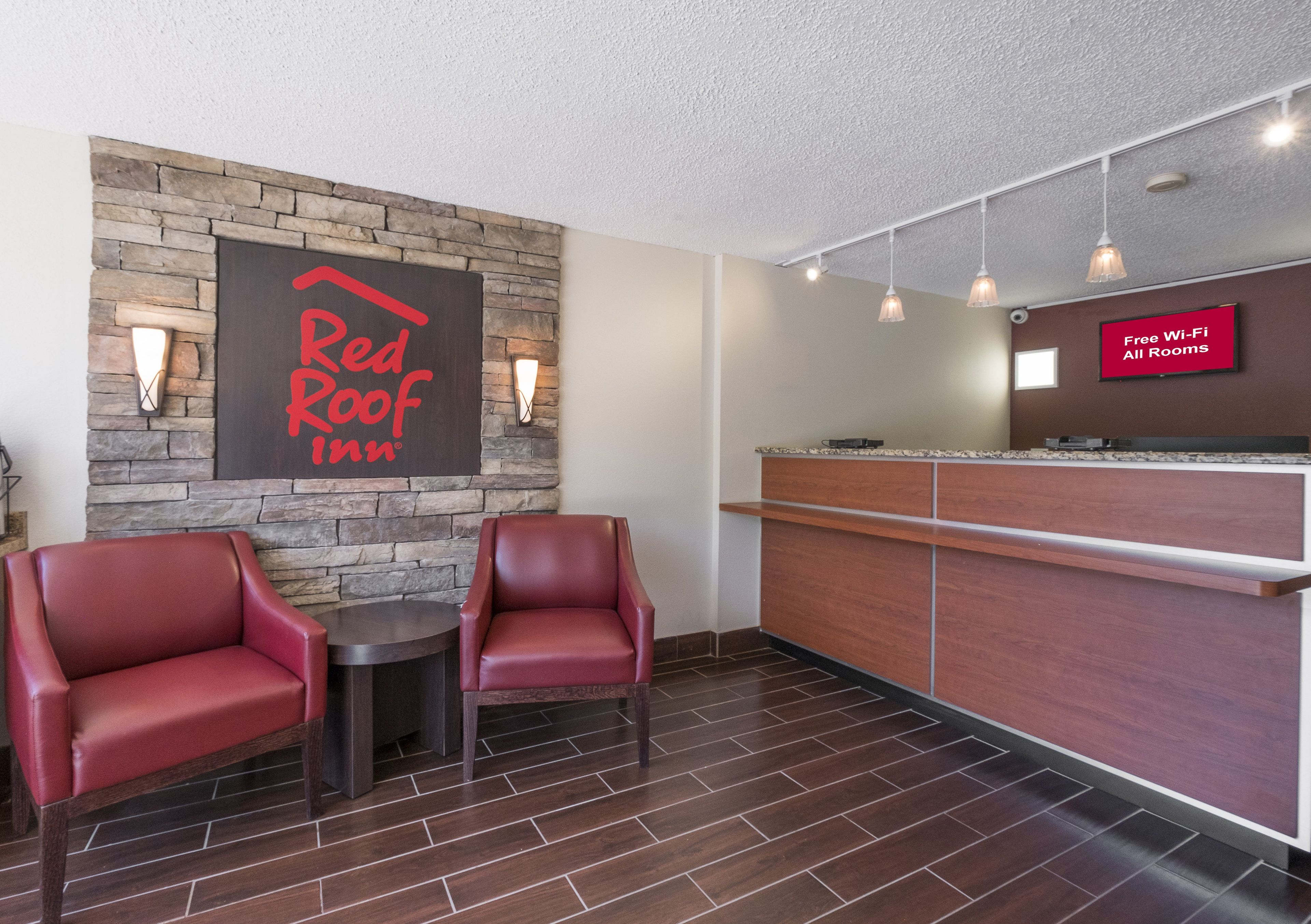 A Red Roof Inn reception area with a stone wall, two leather chairs, a wooden reception desk, and a sign indicating free Wi-Fi in all rooms.