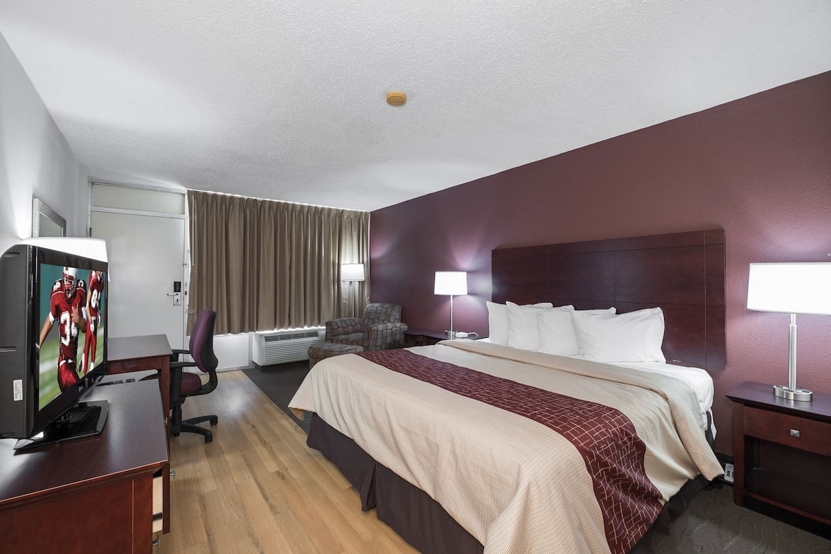 Une chambre d’hôtel avec un lit, un bureau avec une télévision, une chaise, une lampe et une fenêtre avec des rideaux.