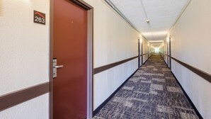 A hotel corridor with a carpeted floor, a door with a number sign, and a hallway leading to a bright end. - Red Roof Inn Dumfries - Quantico (Dumfries)