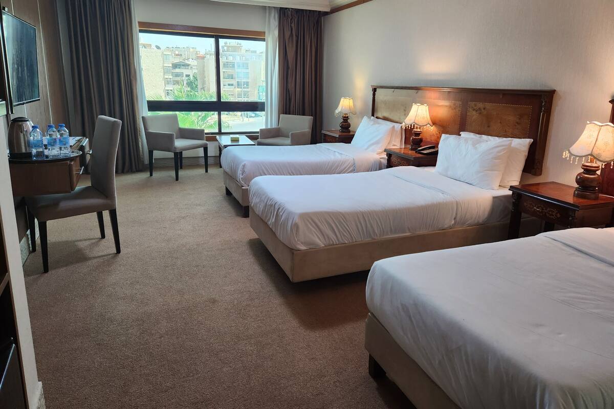 A hotel room with two beds, a desk with a chair, a TV, and a view of buildings outside.