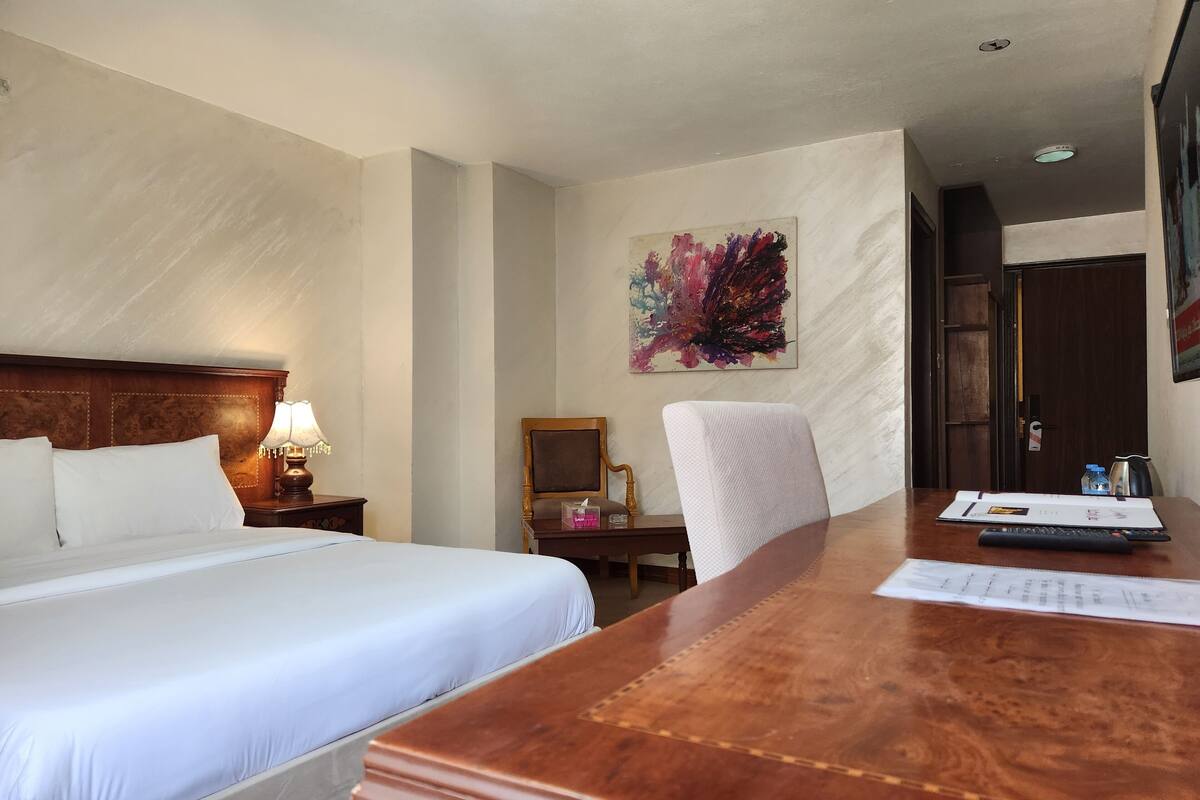 A hotel room with a large bed, a wooden desk, a chair, a desk lamp, a painting on the wall, and a door.