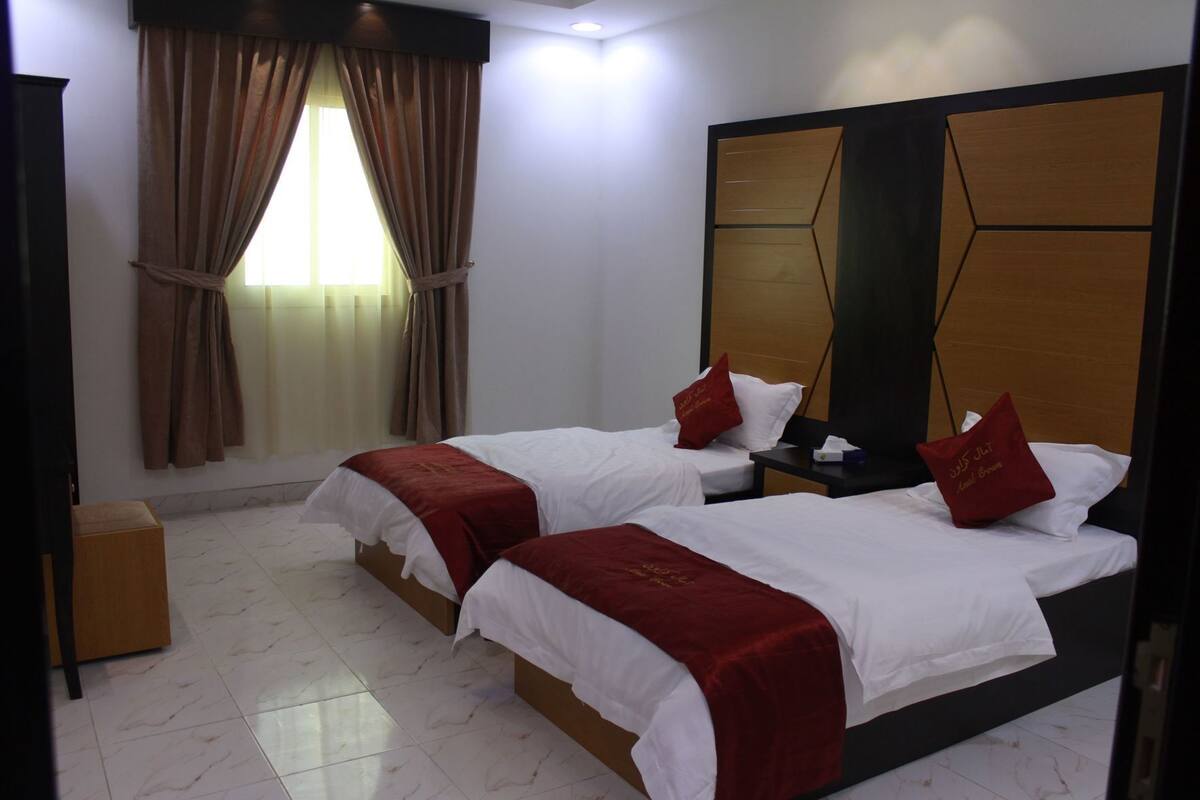 A hotel room with two beds, a wooden headboard, a nightstand, a mirror, and a window with curtains.