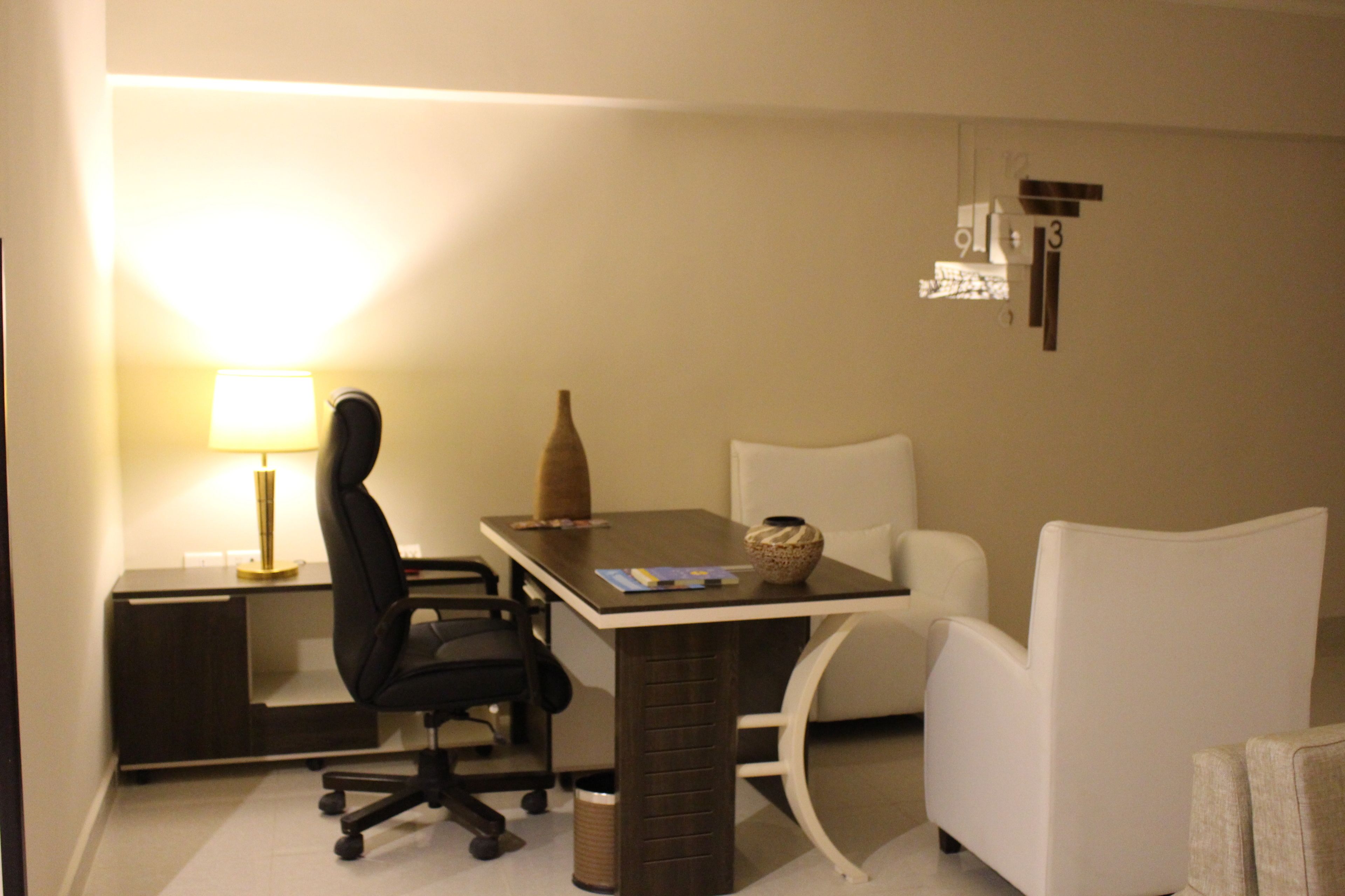 Una habitación de hotel con un escritorio, una silla de oficina, una lámpara y dos sillones blancos.