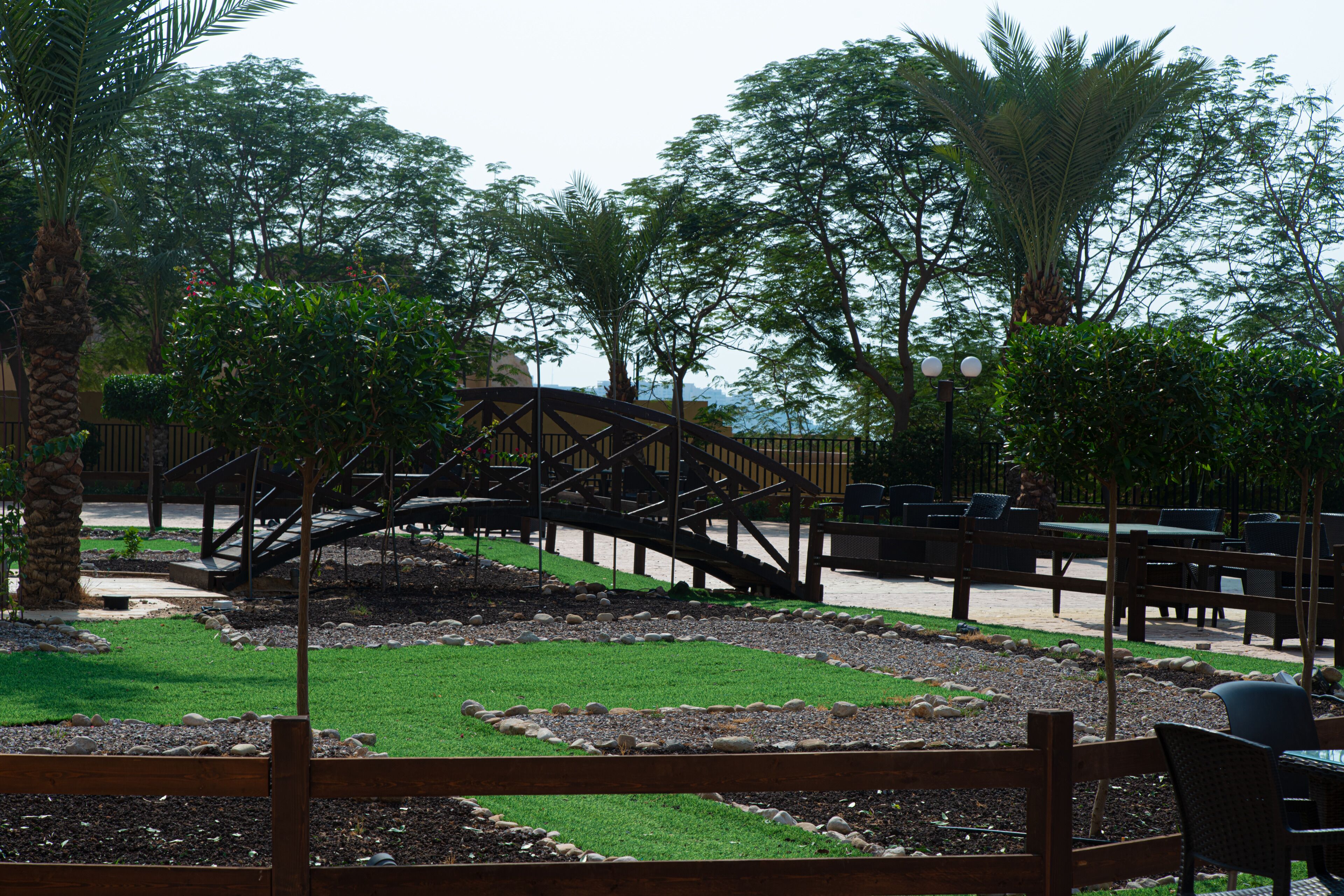 A wooden bridge over a landscaped area with benches and tables, surrounded by trees and a clear sky.