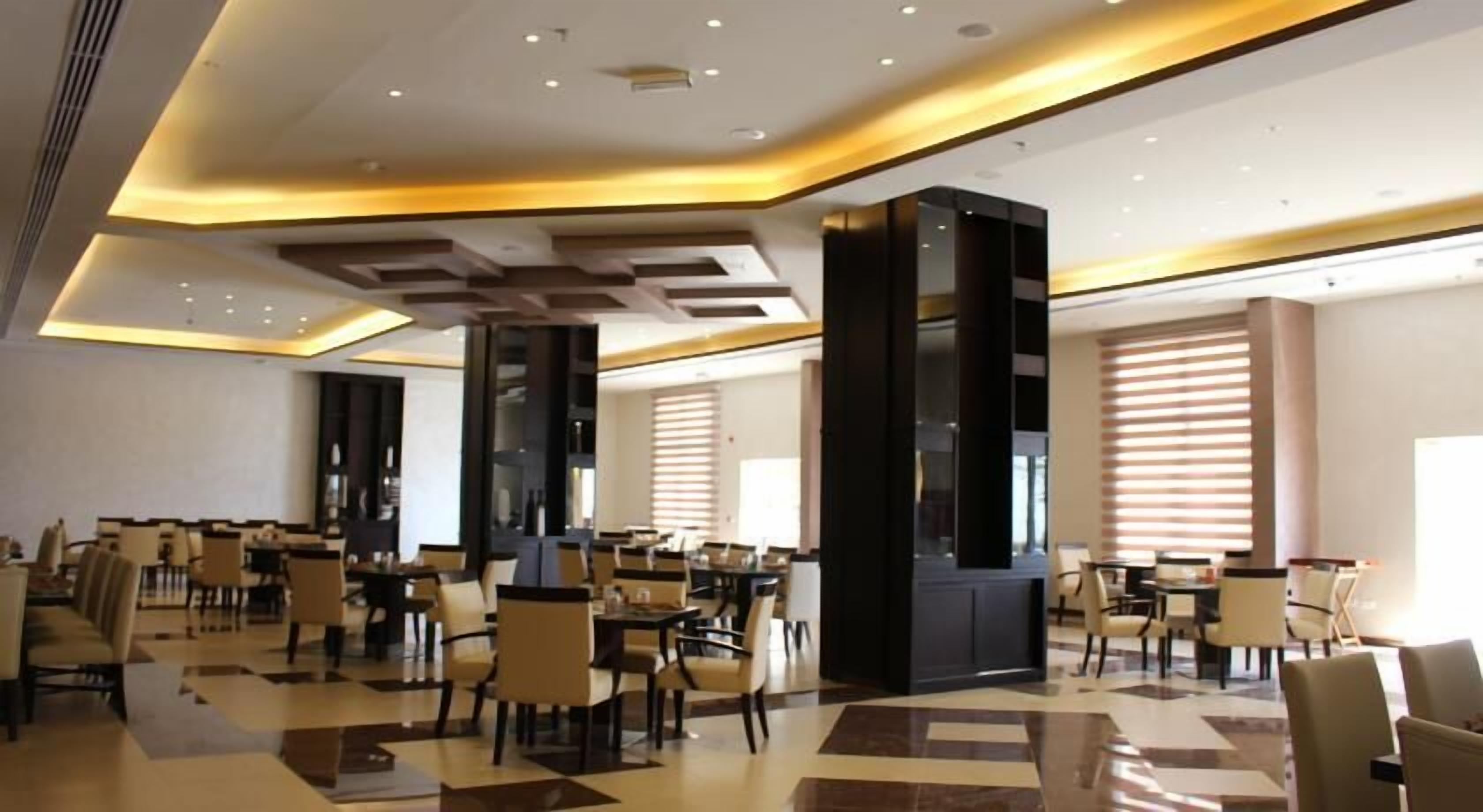 Un amplio vestíbulo de hotel con varias zonas de estar, una columna central y ventanas grandes con persianas.