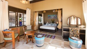 A bedroom with a bed, bedside tables, a chair, a table, and a window with curtains. - Villas des Alizes (Praslin Island)
