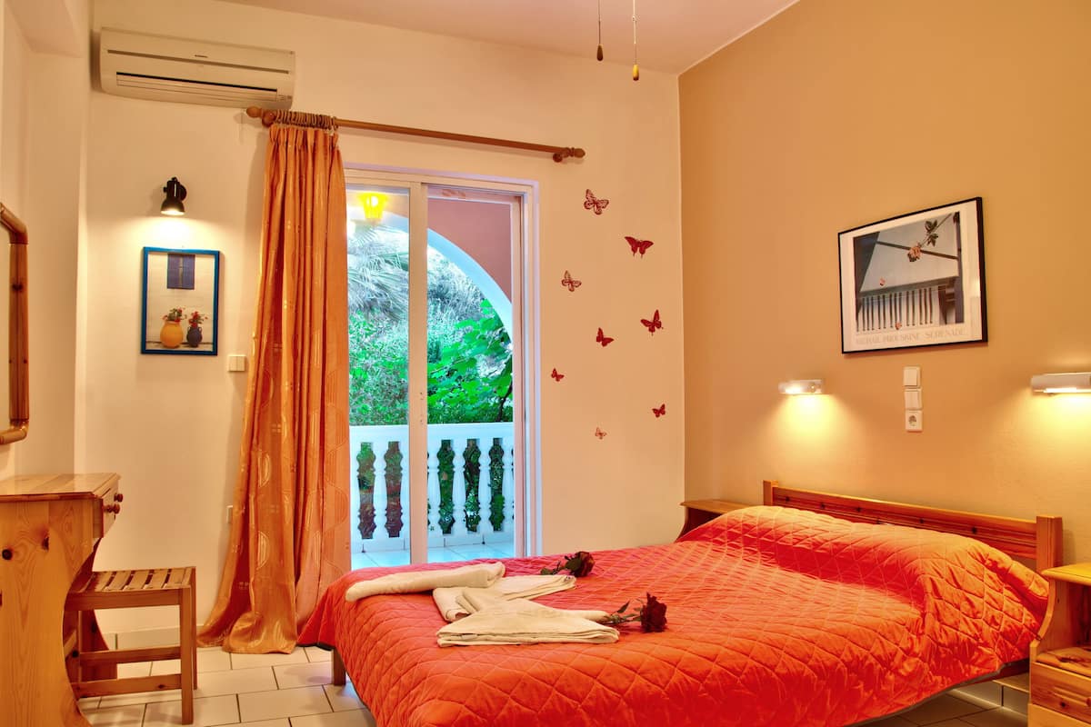 A bedroom with a large bed, a wooden desk, a chair, a window with curtains, and a balcony with a railing.