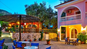An outdoor patio with round tables and blue chairs, a stone wall, and a building with a balcony. - Kyprianos (Zakynthos)