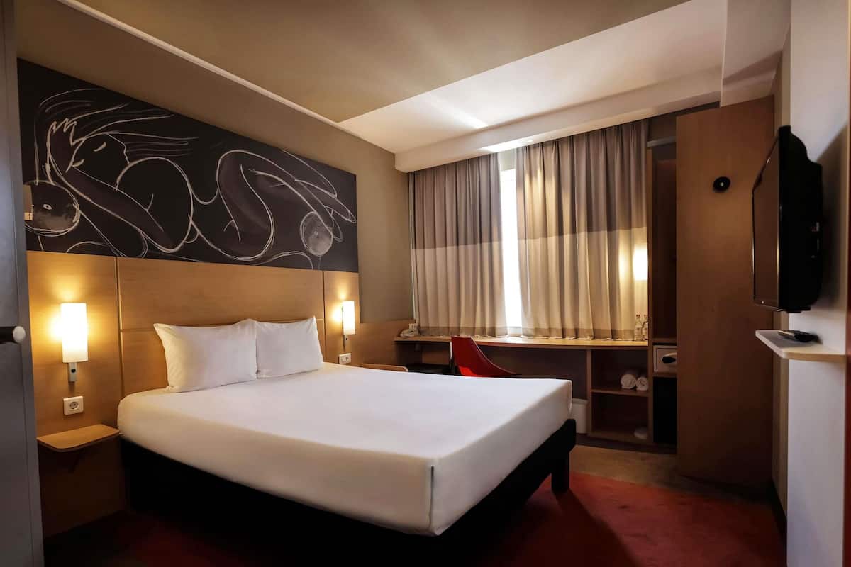 A hotel room with a large bed, two bedside lamps, a desk with a chair, a television, and a window with curtains.