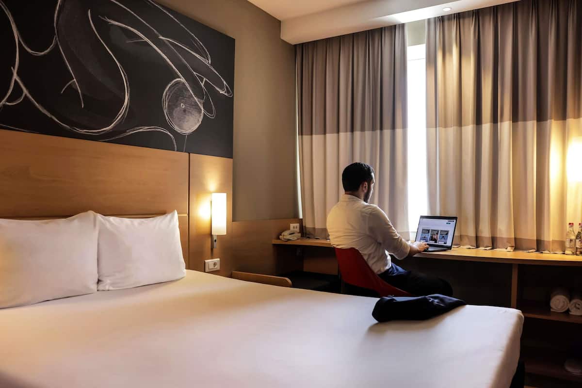 A hotel room with a large bed, a desk, a laptop, and a wall-mounted light.