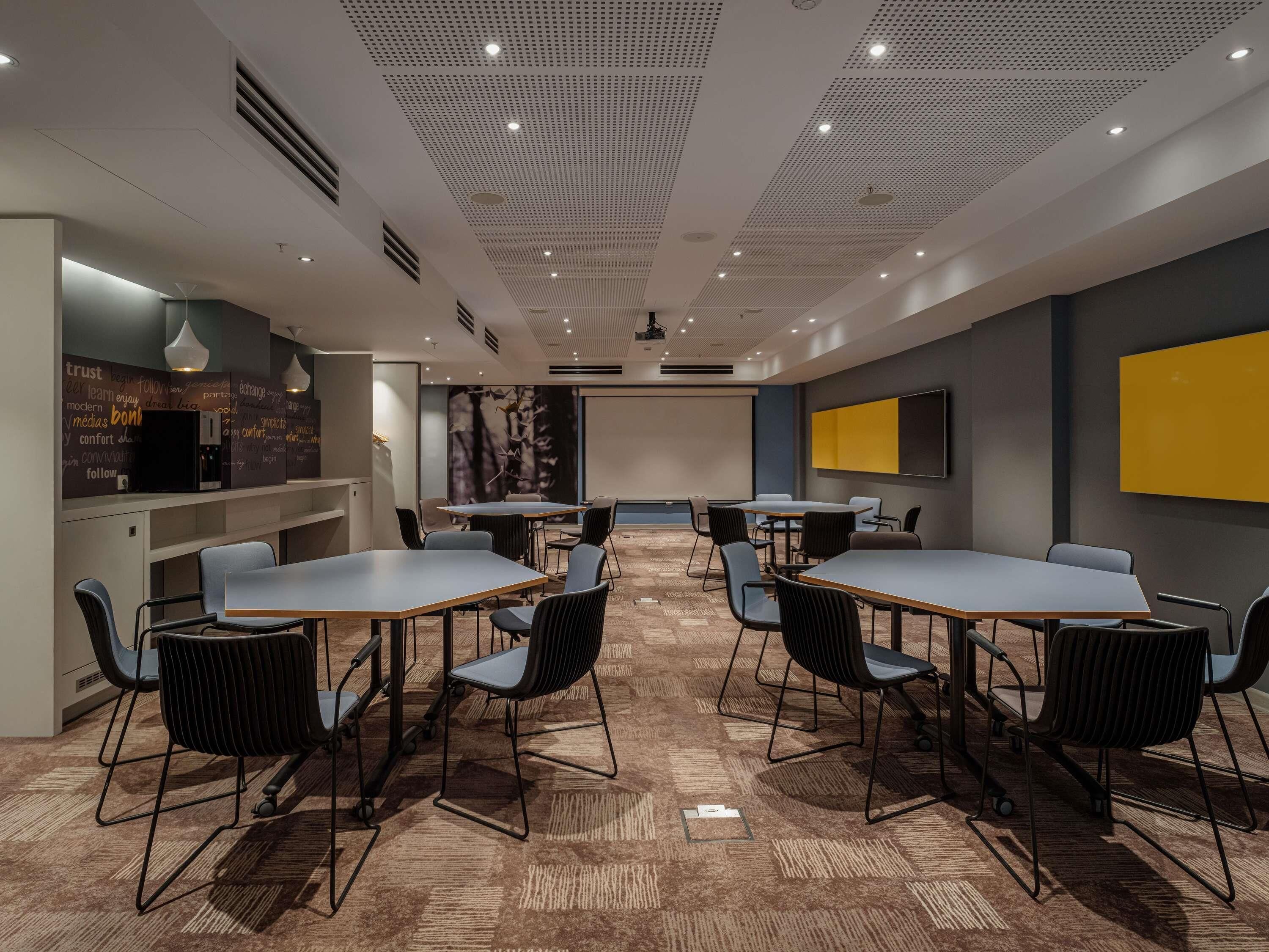 A conference room with tables, chairs, a projector screen, and a large yellow wall.