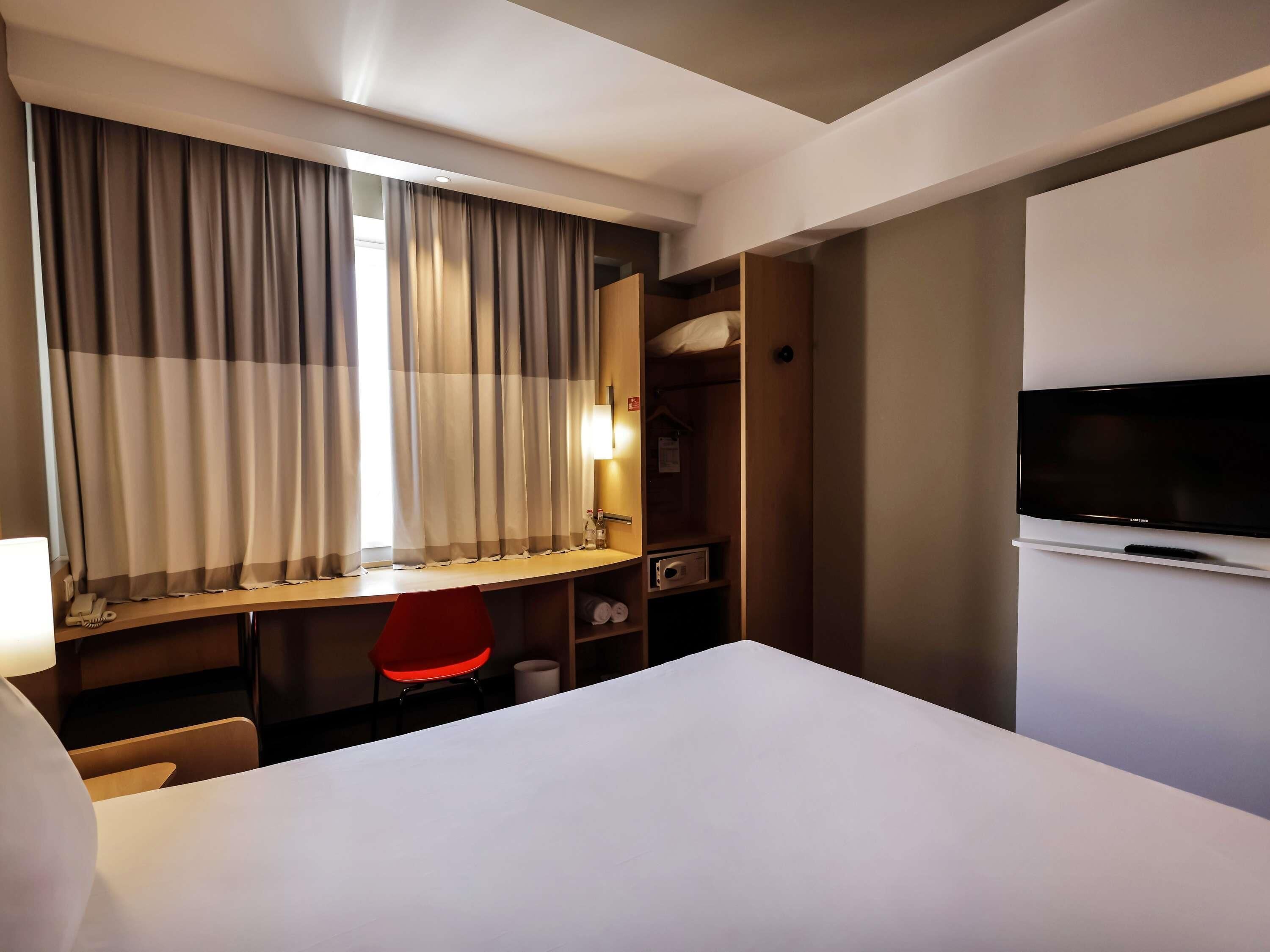 A hotel room with a bed, a desk with a chair, a television, and a wardrobe.