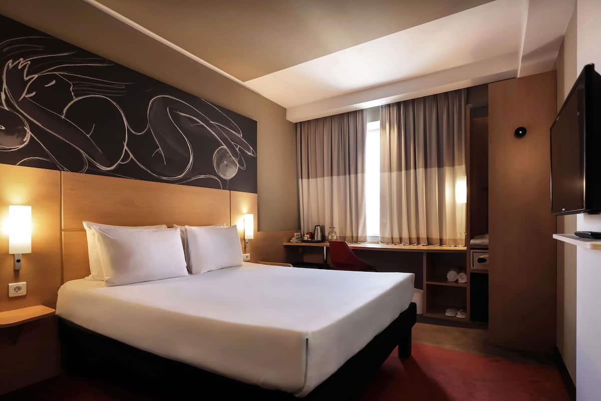 A hotel room with a large bed, a desk, a chair, a TV, and a wall art piece.