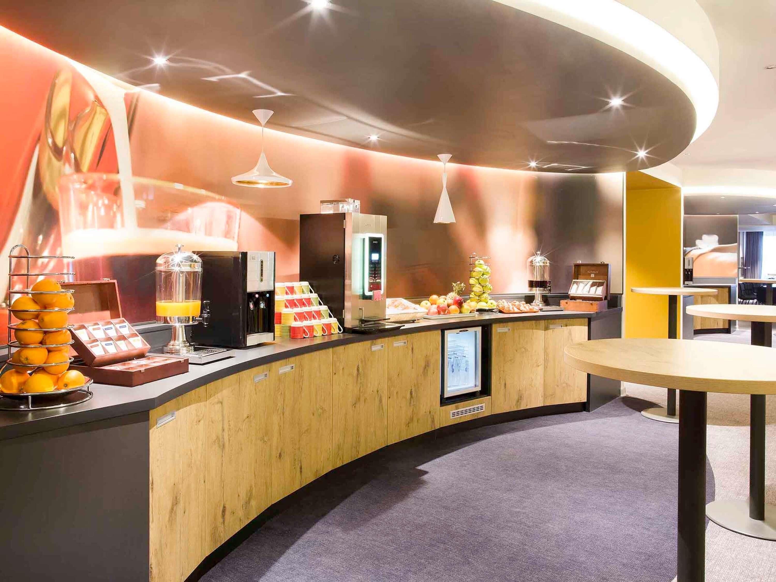 Ein modernes Hotel-Foyer mit einer gebogenen Theke, einer Kaffeemaschine, einer Obstauslage und einem digitalen Bildschirm.