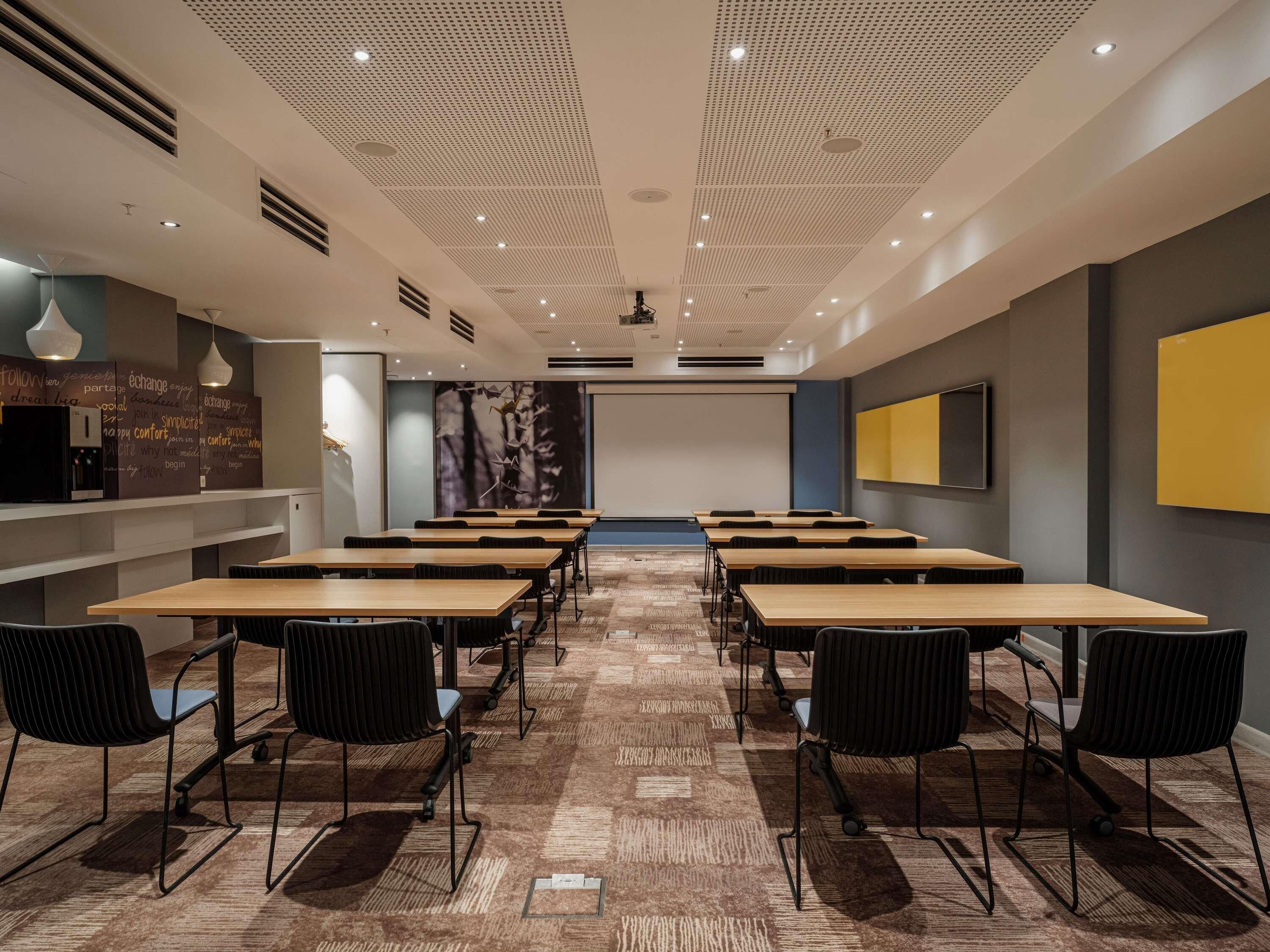 A conference room with multiple tables and chairs, a projector screen, and a large wall with a screen and wall art.