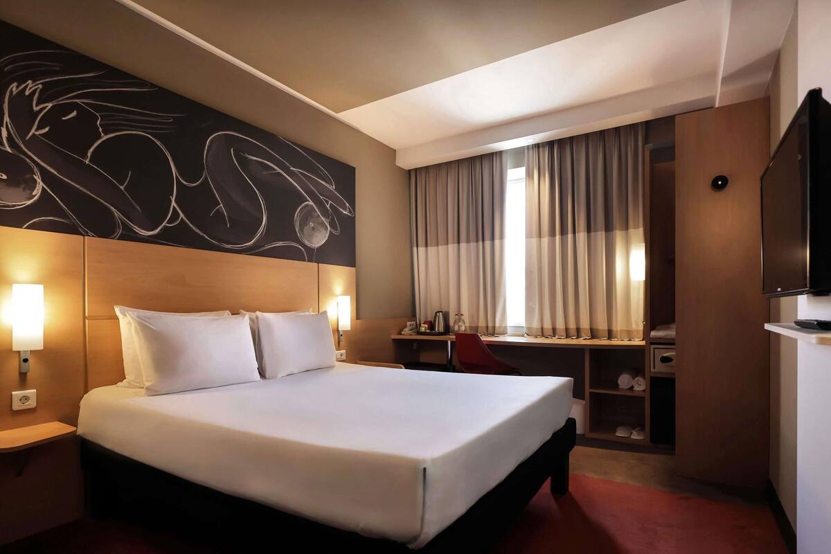 Una habitación de hotel con una cama grande, un escritorio, una silla, un televisor y una obra de arte en la pared.
