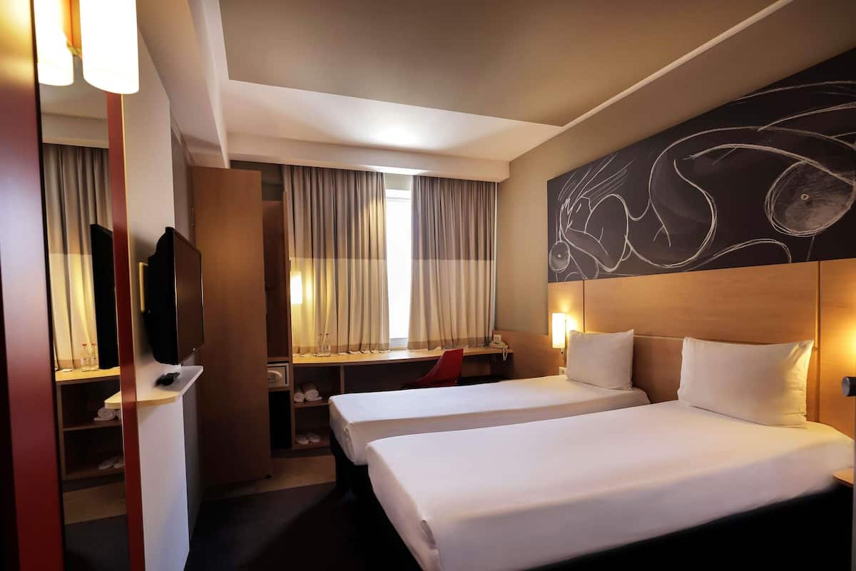 A hotel room with two beds, a desk with a chair, a television, and a large abstract painting on the wall.