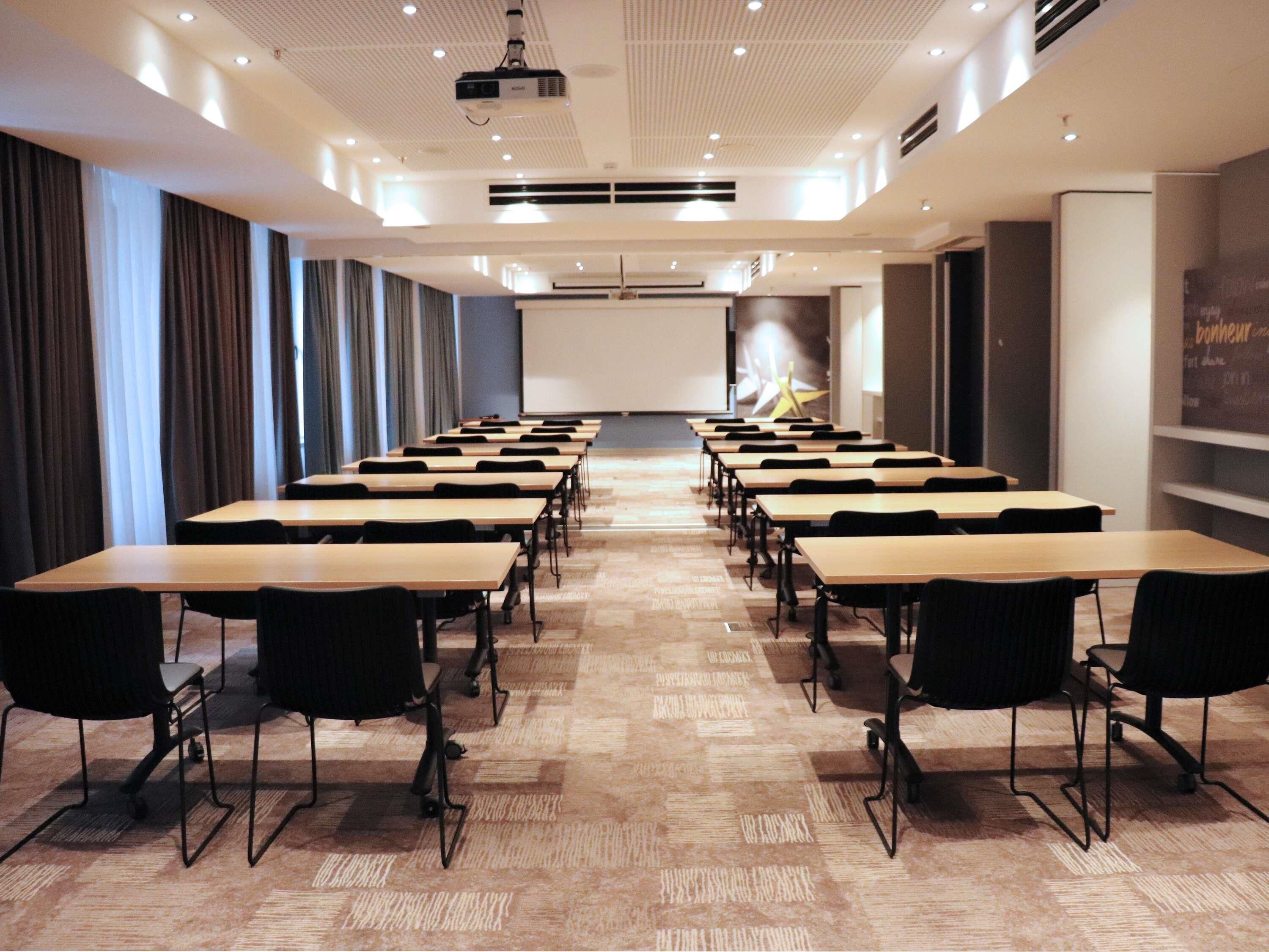A conference room with rows of tables and chairs, a projector screen, and a large screen on the wall.