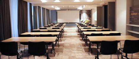 Une salle de conférence avec des rangées de tables et de chaises, un écran de projection et un grand écran mural.