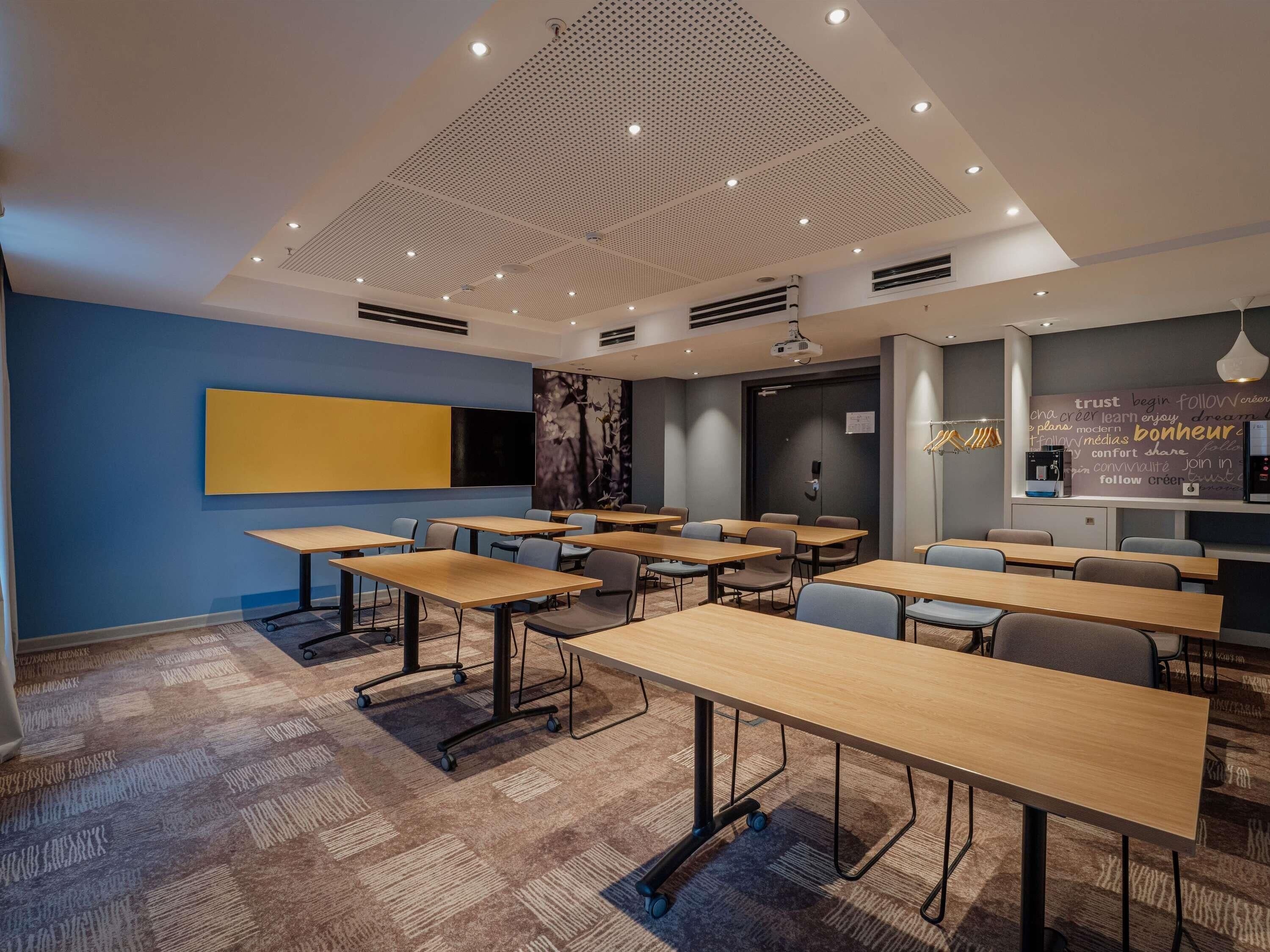 Un salón de conferencias moderno con mesas, sillas, un proyector y una pared con mensajes motivacionales.