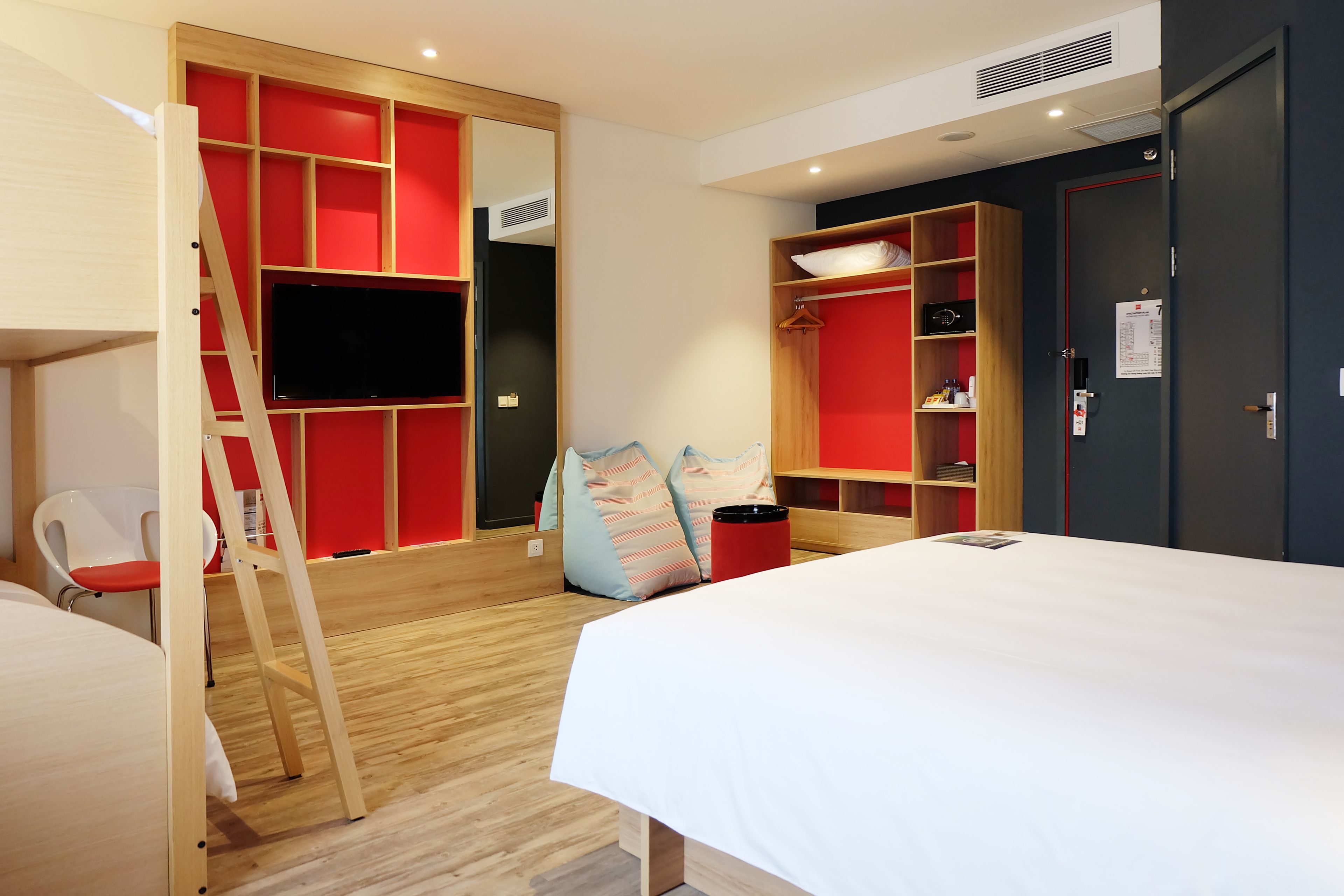 A modern hotel room with a bed, a TV mounted on a red wall, a wardrobe, and a wooden ladder.