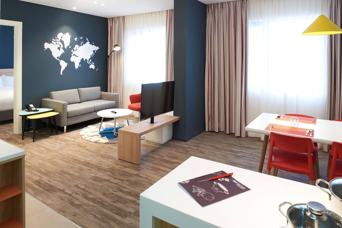 Habitación de hotel moderna con sofá, televisión, mesa de comedor con sillas, piso de madera y un mapa mundial en la pared.