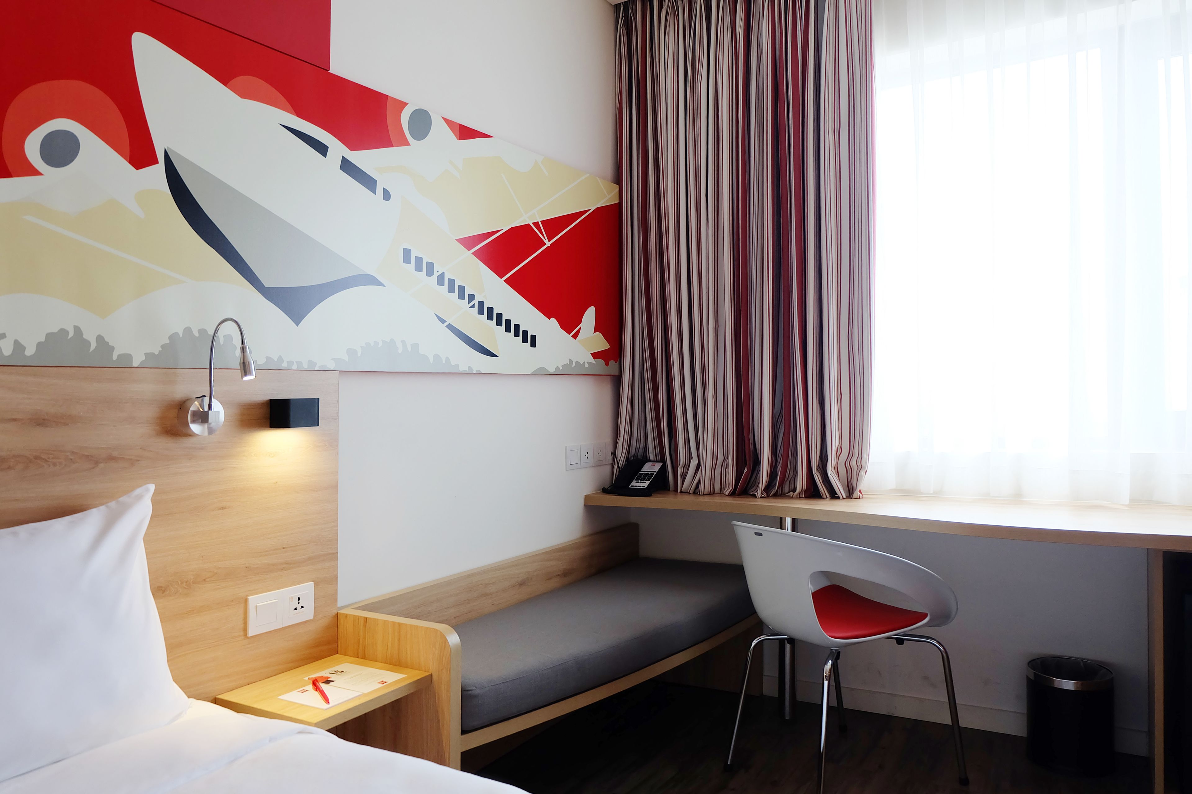 A hotel room with a bed, a desk with a chair, a small bedside table, a wall-mounted telephone, and a window with curtains.