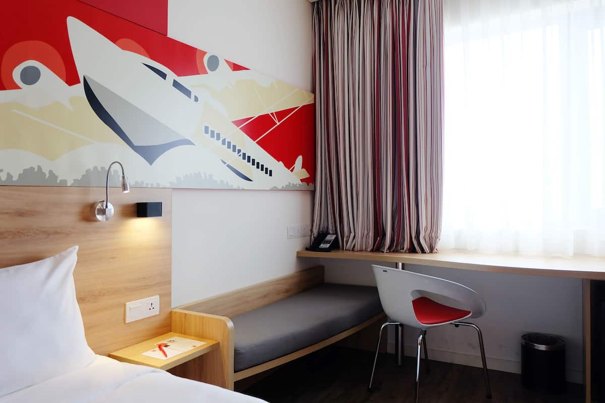 Una habitación de hotel con cama, escritorio y silla, mesita de noche, teléfono en la pared y ventana con cortinas.