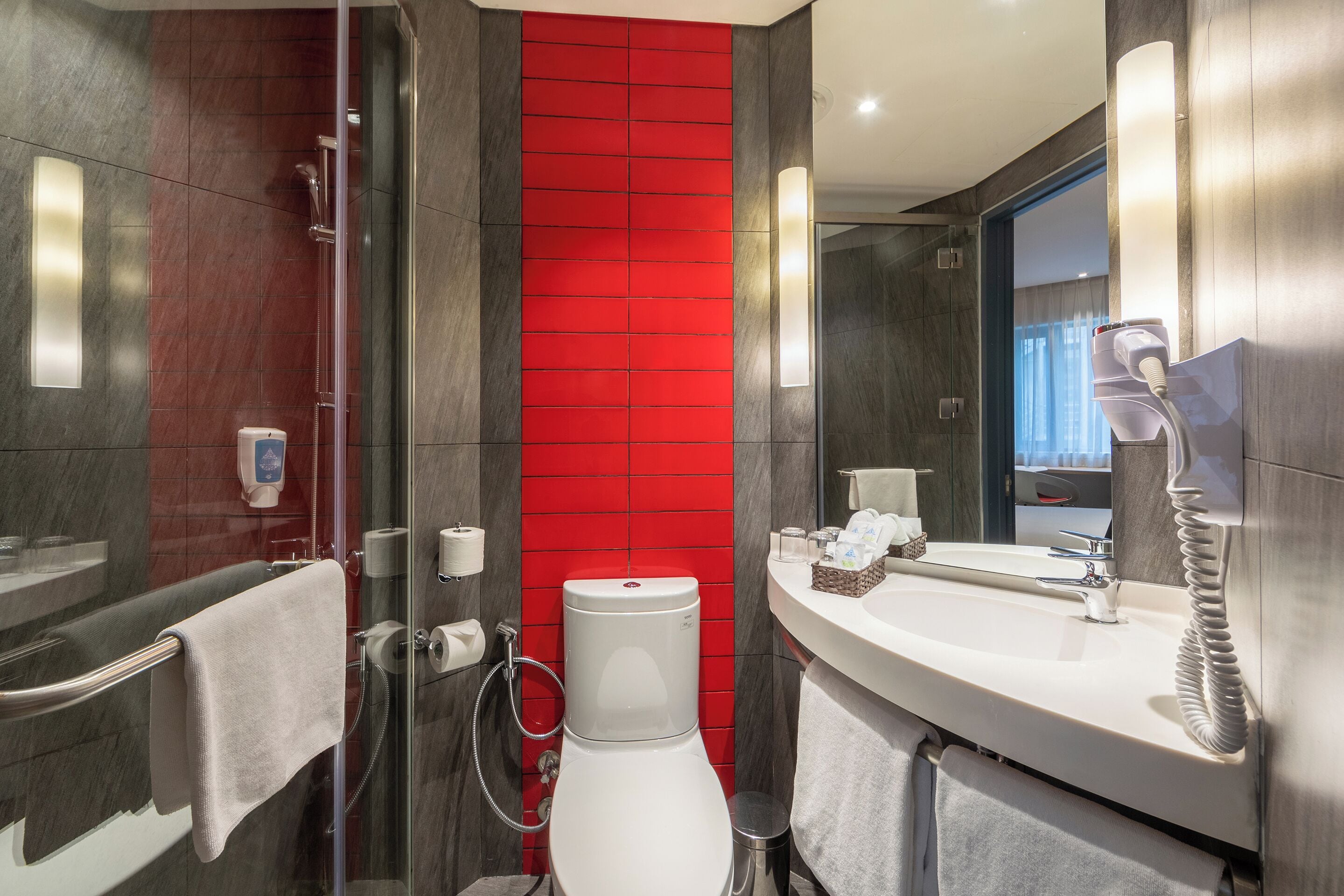 A modern bathroom with a glass shower enclosure, a white toilet, a sink with a mirror, and a red accent wall.