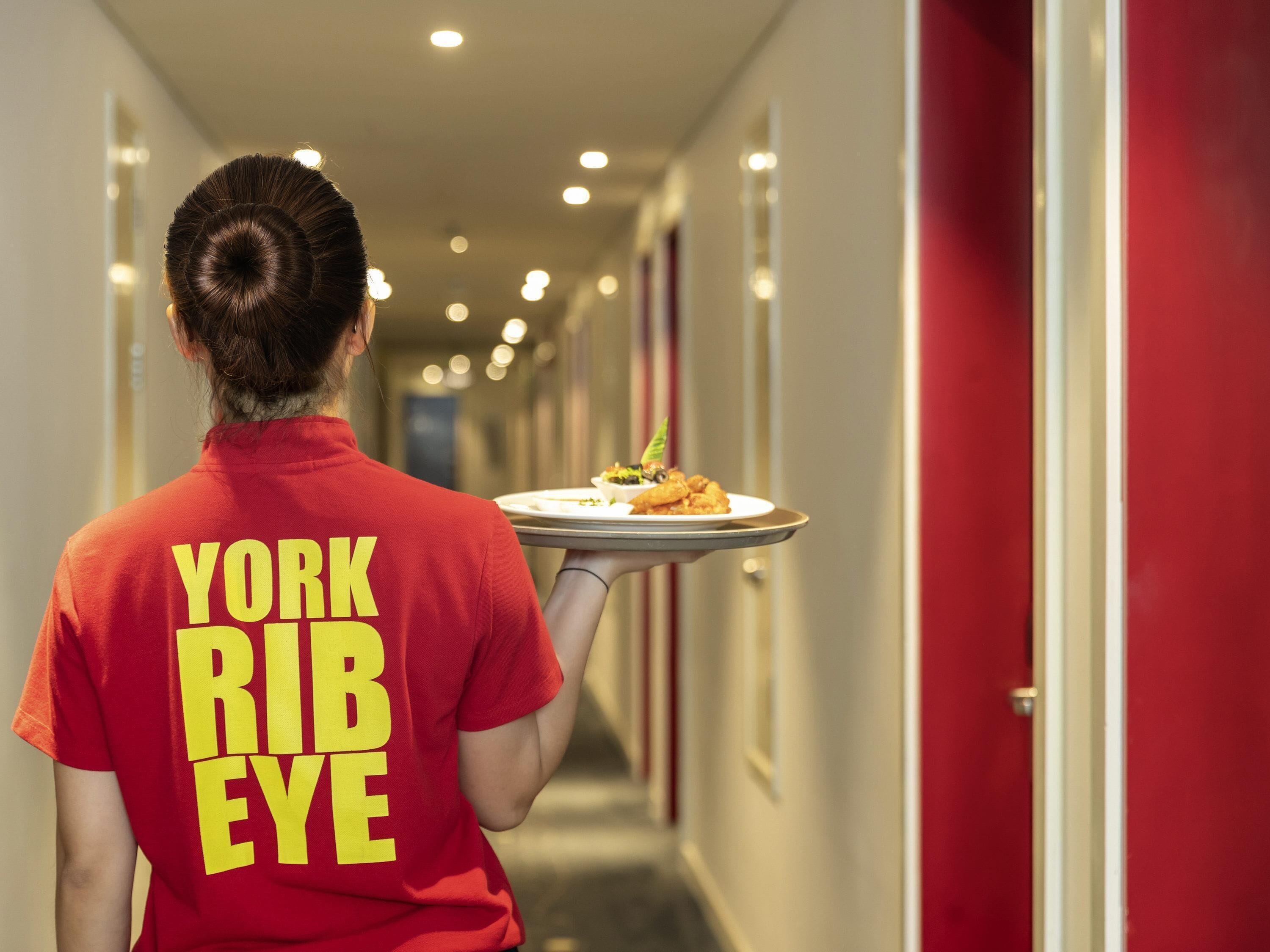 A person in a red shirt with 'YORK RIB BEE' text walking down a hotel corridor carrying a tray with food.