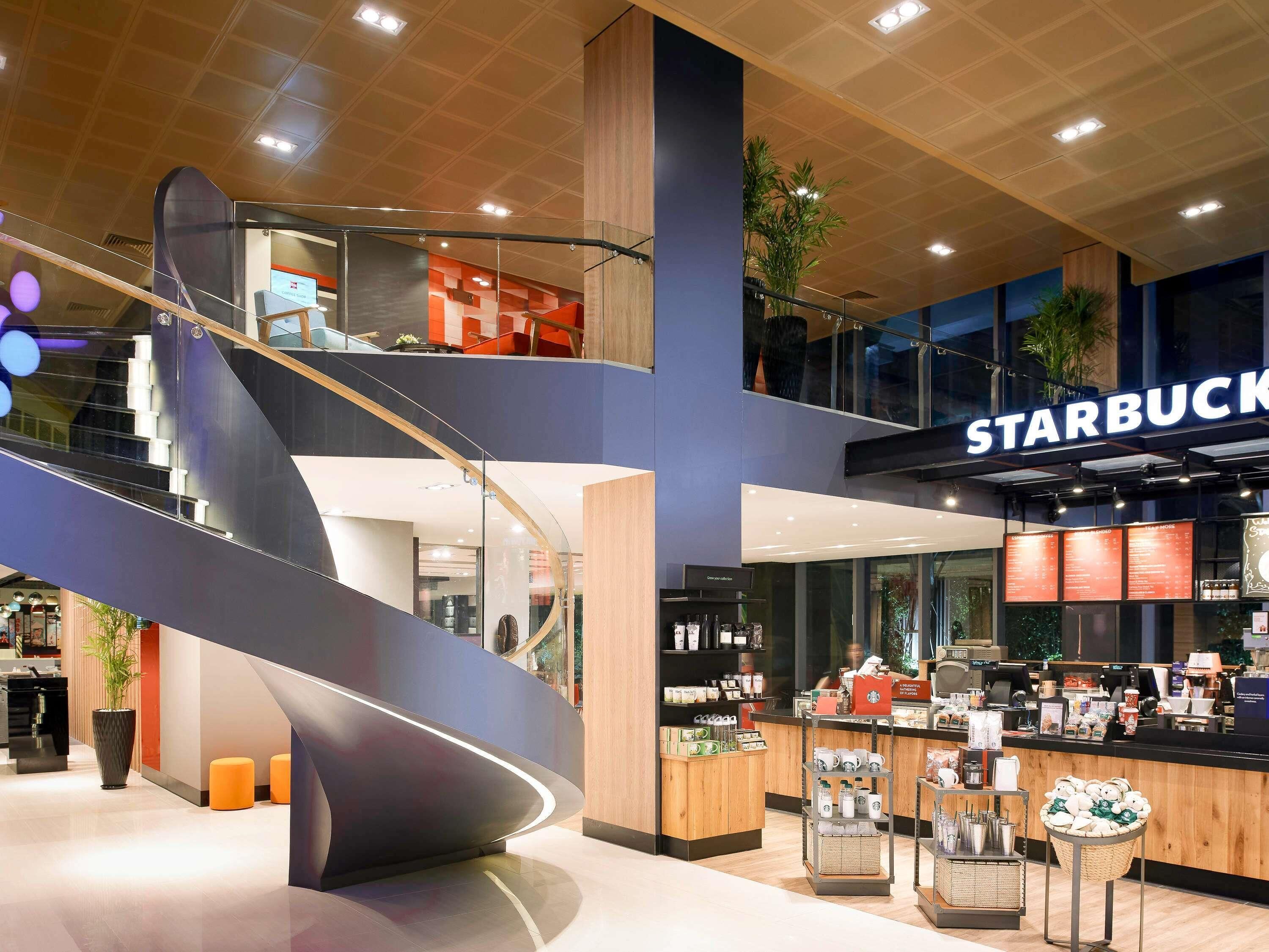 Un interior moderno con una cafetería Starbucks, una escalera curva y un área de descanso con taburetes naranjas.