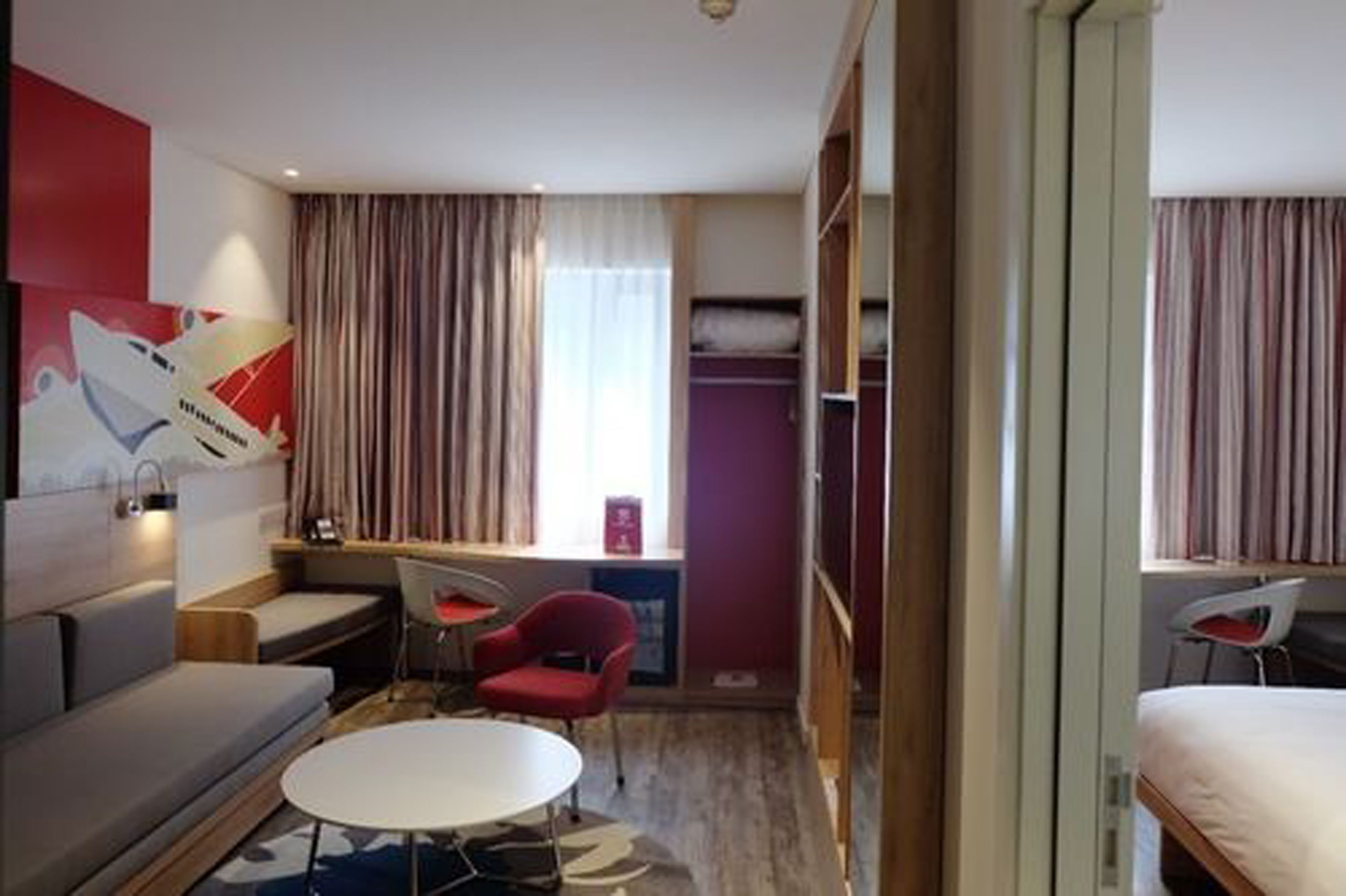 A modern hotel room with a sofa, a small round table, a red chair, a bed, and a wardrobe.