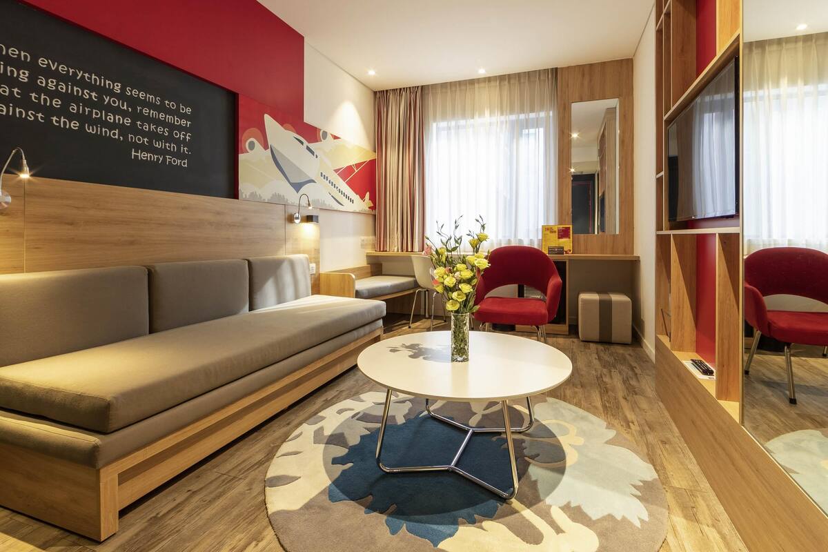 Una habitación de hotel moderna con un sofá, una mesa redonda, una silla roja, un espejo y una pared con una cita.