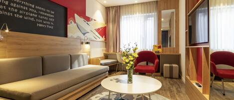A modern hotel room with a sofa, a round table, a red chair, a mirror, and a wall with a quote.
