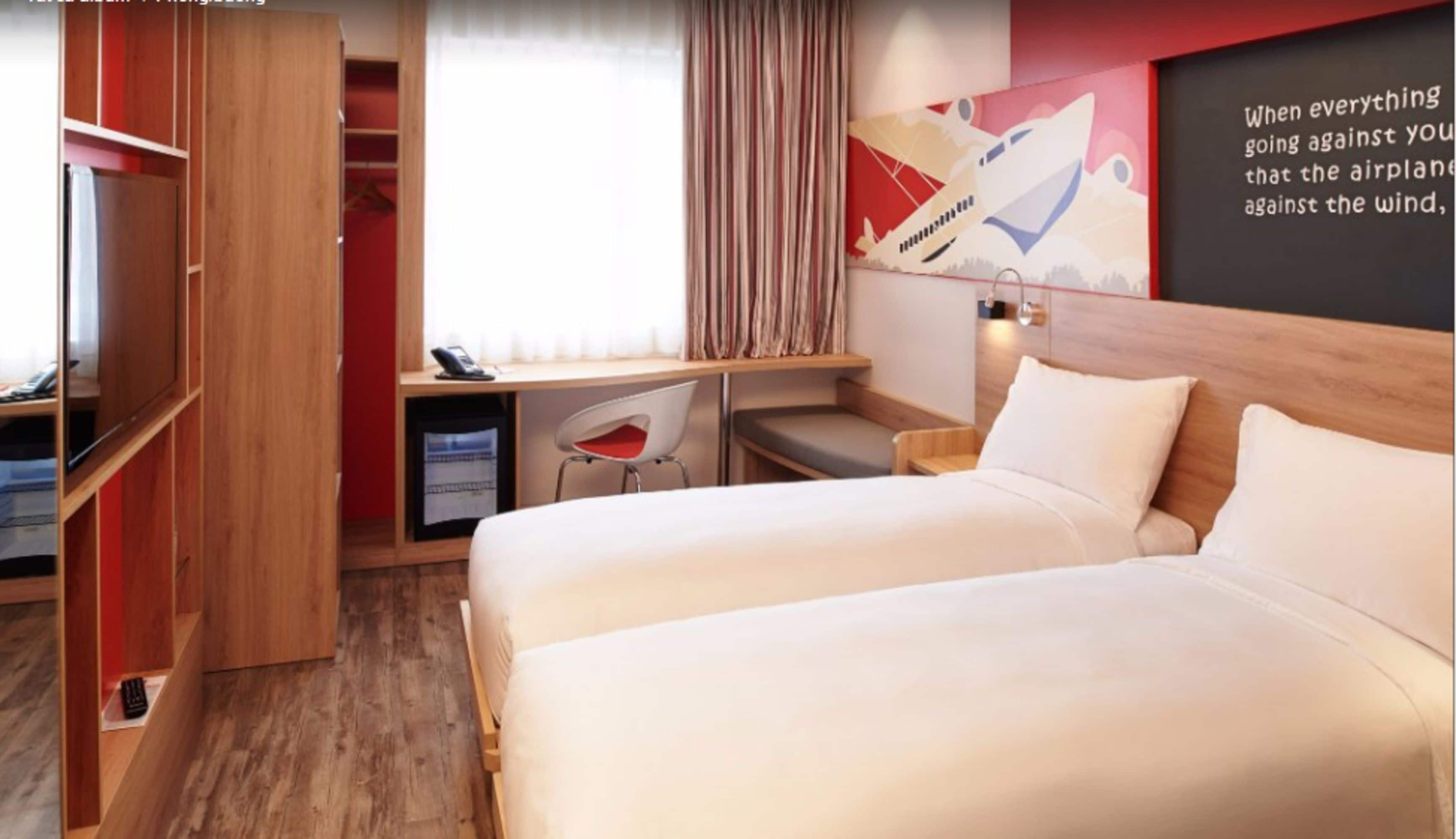 A hotel room with a bed, a desk, a chair, a wardrobe, and a wall with a motivational quote.