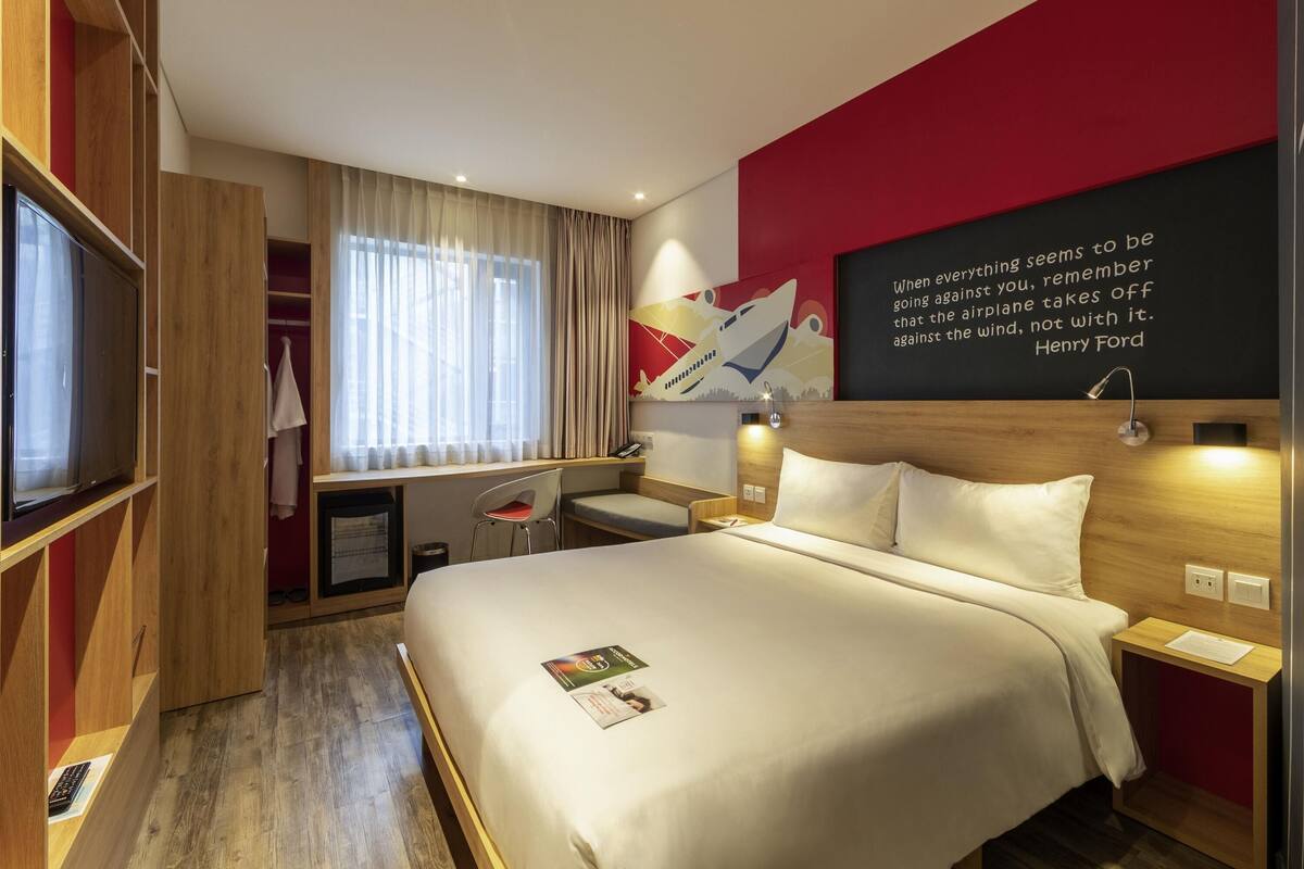 Una habitación de hotel con cama, escritorio, silla, televisión y una pared con una cita.