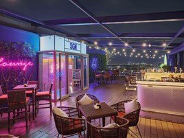 Een terras met buitenbar, rieten meubels, een neonbord en uitzicht over de stad.