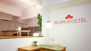 Una camera moderna e minimalista con una scrivania in legno, una pianta in vaso e un cartello a parete che recita 'PLUM HOSTEL'.