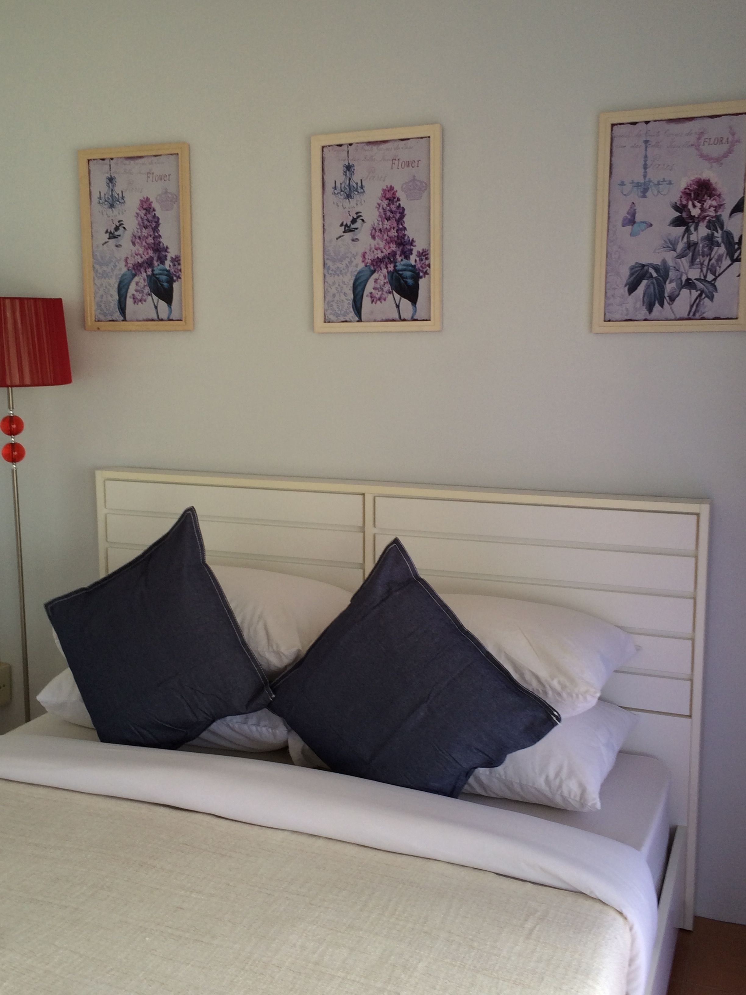 Un dormitorio con cama, almohadas, una mesita de noche y una lámpara. Hay tres cuadros con motivos florales en la pared.