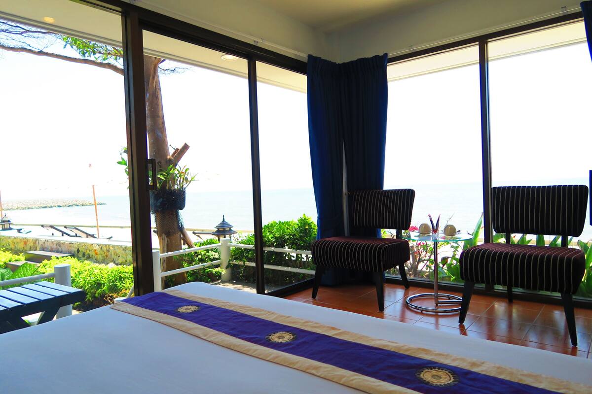 A room with a bed, two striped chairs, a small table with a lamp, and a view of the sea.