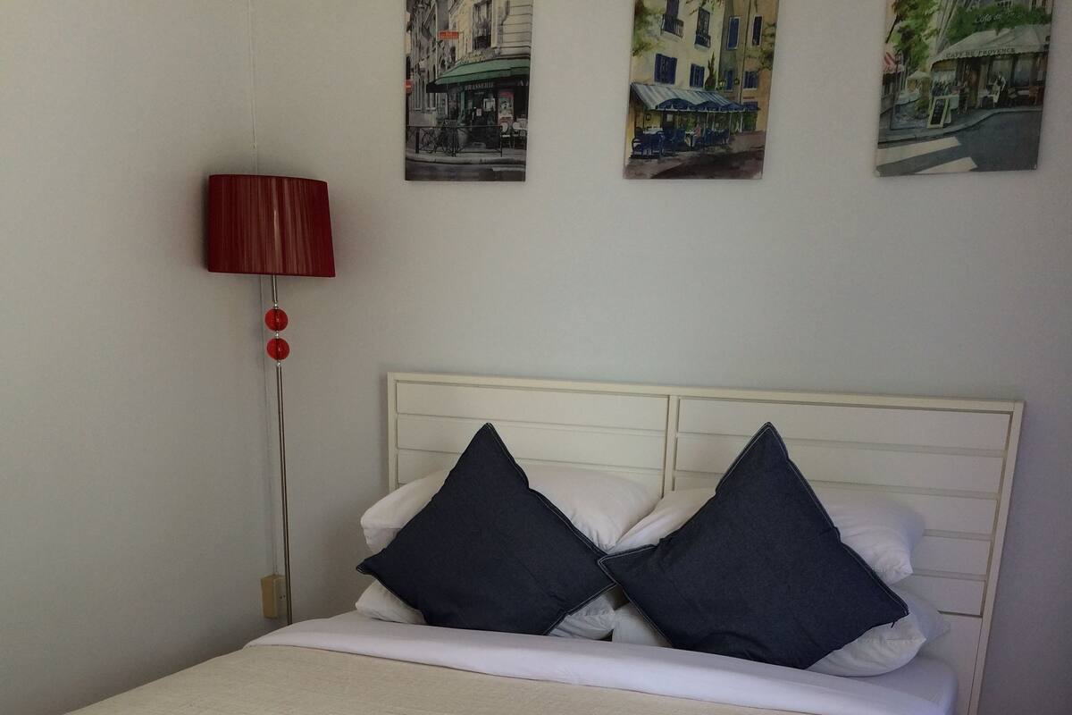 A bed with a white headboard, two dark pillows, and a beige blanket. There is a red lamp on a stand beside the bed, and three framed pictures on the wall above.