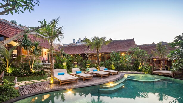 A pool area with lounge chairs and a wooden deck, surrounded by tropical plants and a building with a tiled roof. - Umakayu Bali (Canggu)