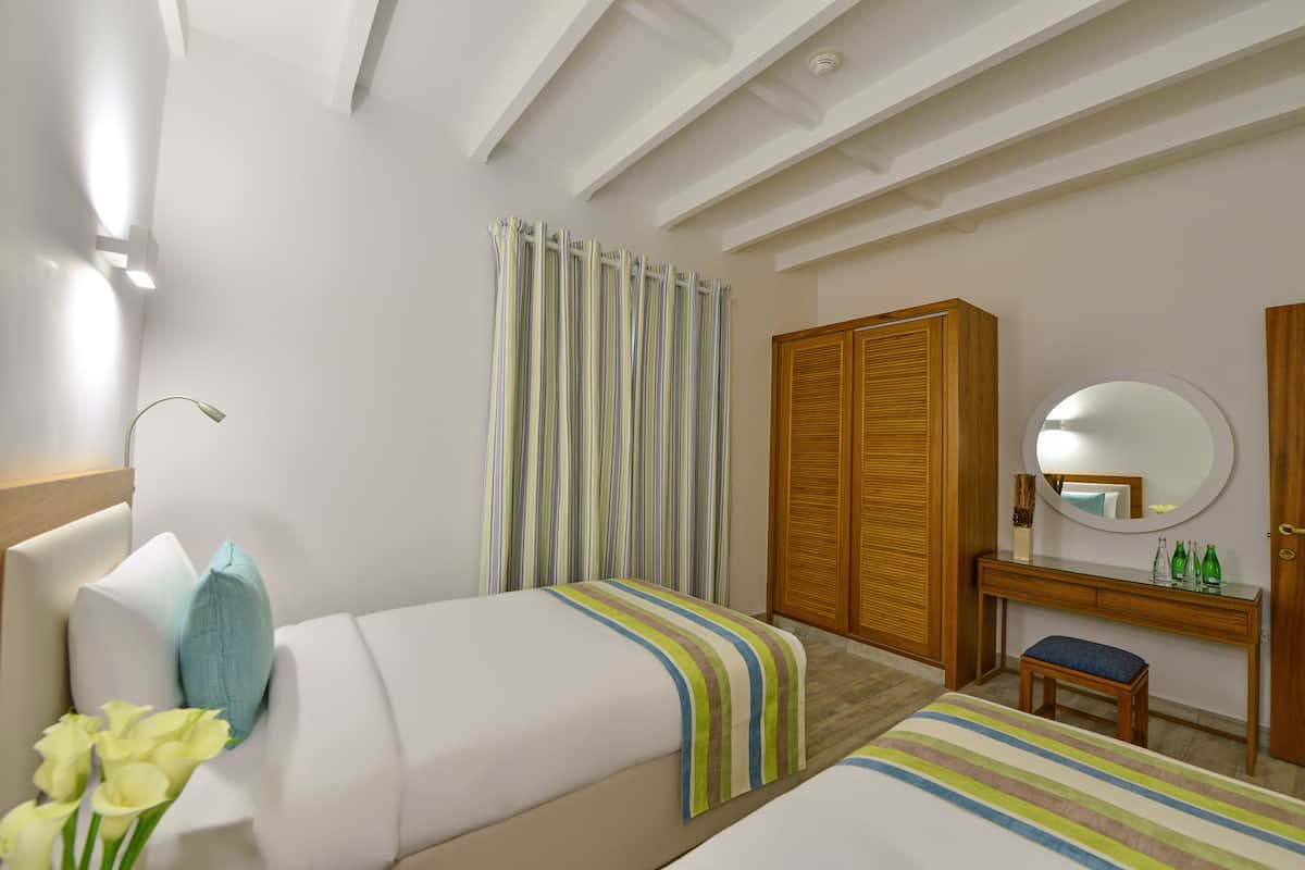 Quarto de hotel com duas camas, um guarda-roupa de madeira, uma toucador com espelho e um banquinho pequeno.