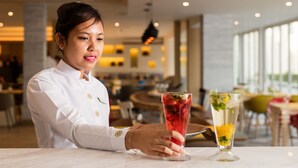 A person in a white uniform holding a tall glass with a layered drink, seated at a table in a well-lit indoor setting with modern decor.