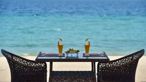 A table with two drinks and a fruit bowl on a beach overlooking the ocean.