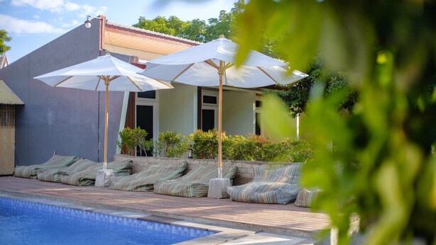 Piscina con sillones de descanso y sombrillas, rodeada de un jardín ajardinado y un edificio con techo de tejas.