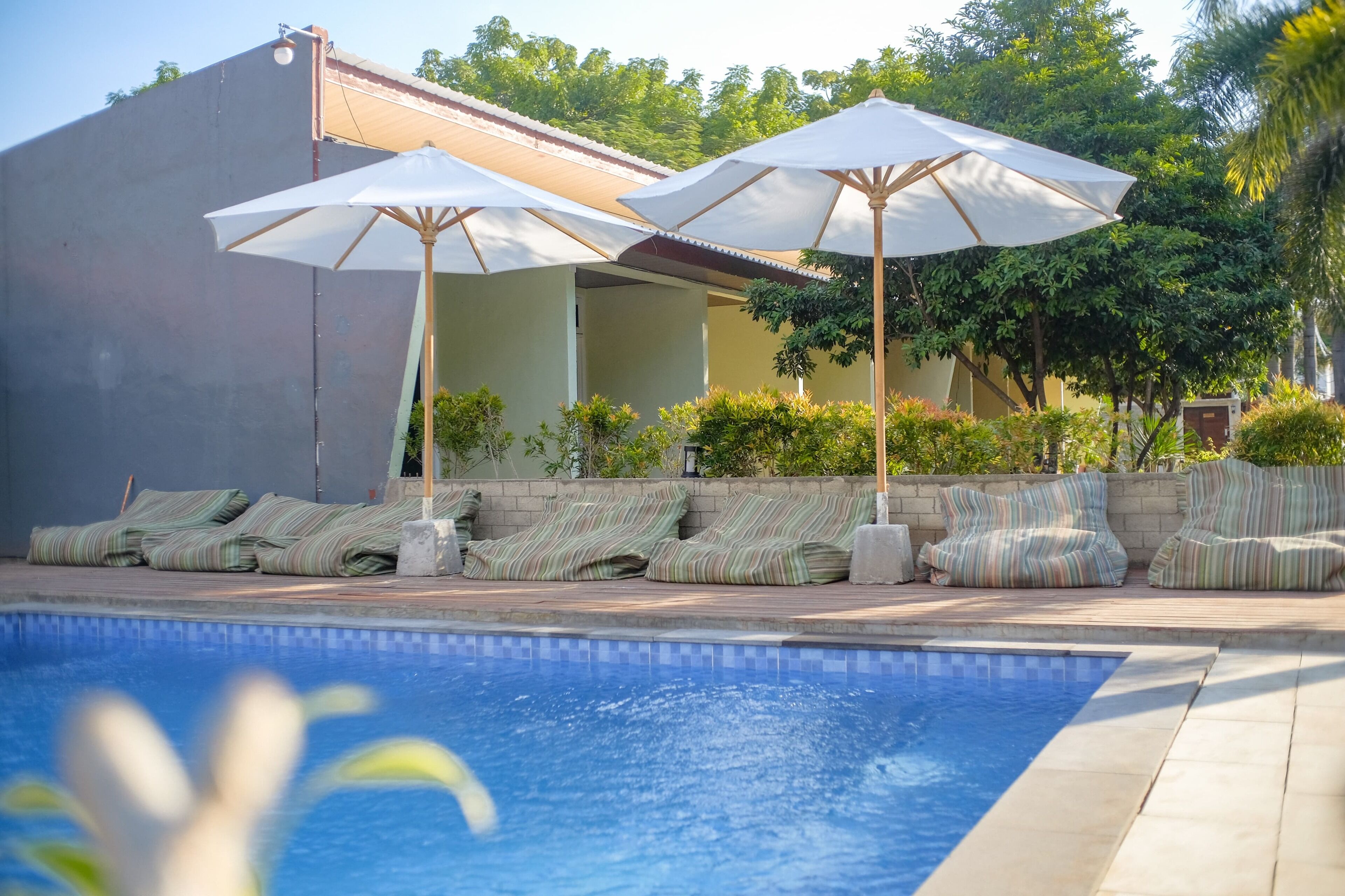 A swimming pool with lounge chairs and umbrellas, surrounded by greenery and a building.